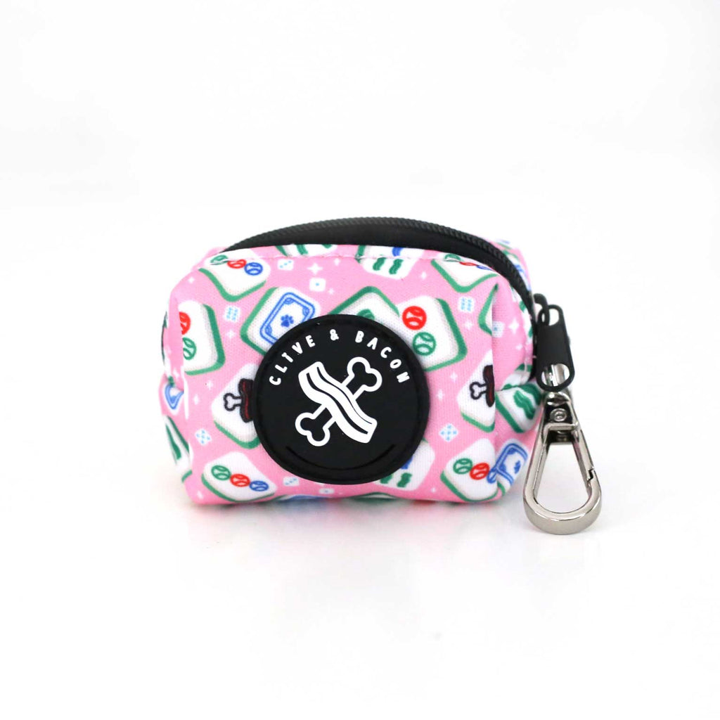 A pink zippered pet waste holder with a colorful classic mahjong tile pattern, featuring a black circular 'CLIVE & BACON' logo patch and a silver metal carabiner clip.