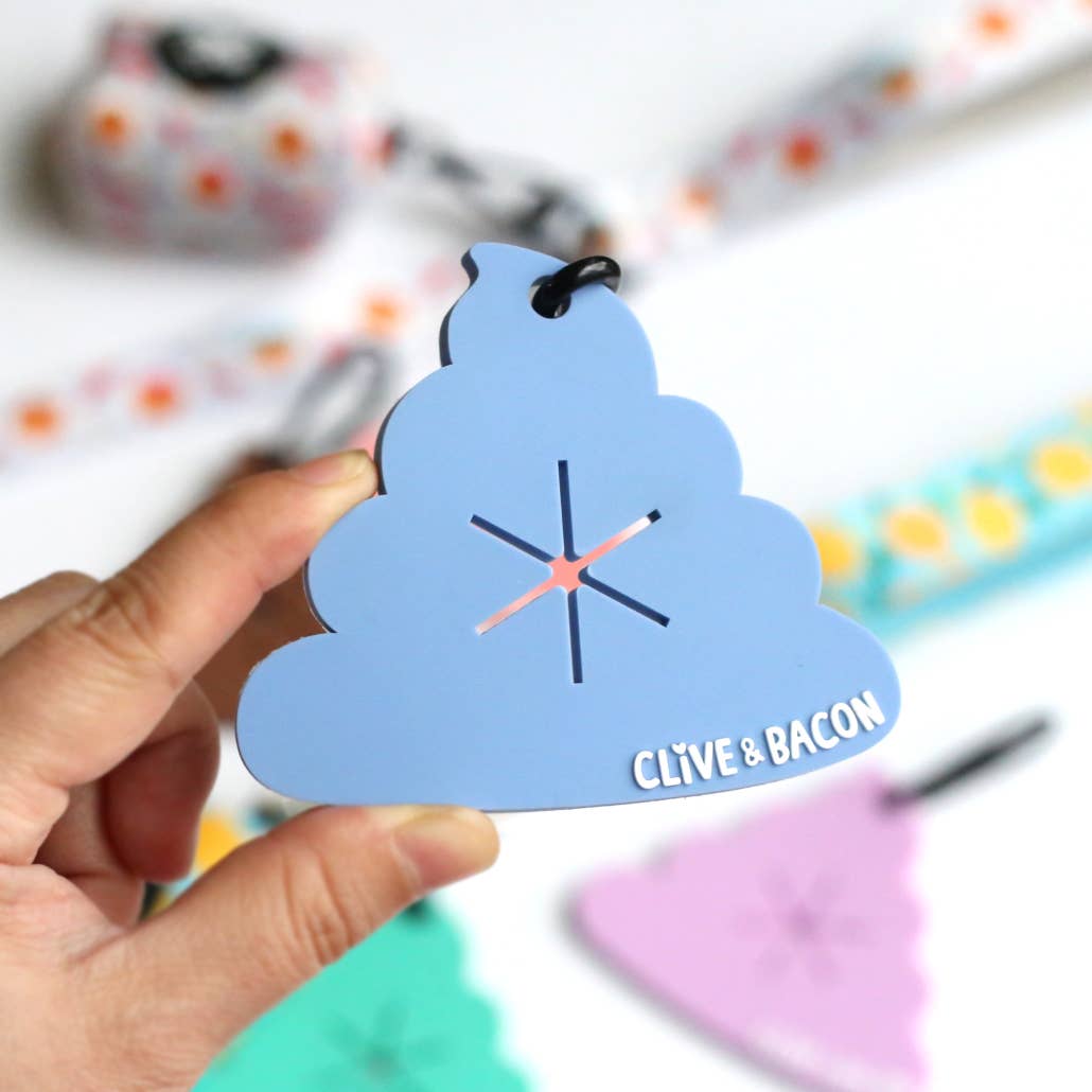 A hand holding a light blue cloud-shaped keychain with a poop symbol and the text 'CLIVE & BACON' in white, with a blurred colorful background.