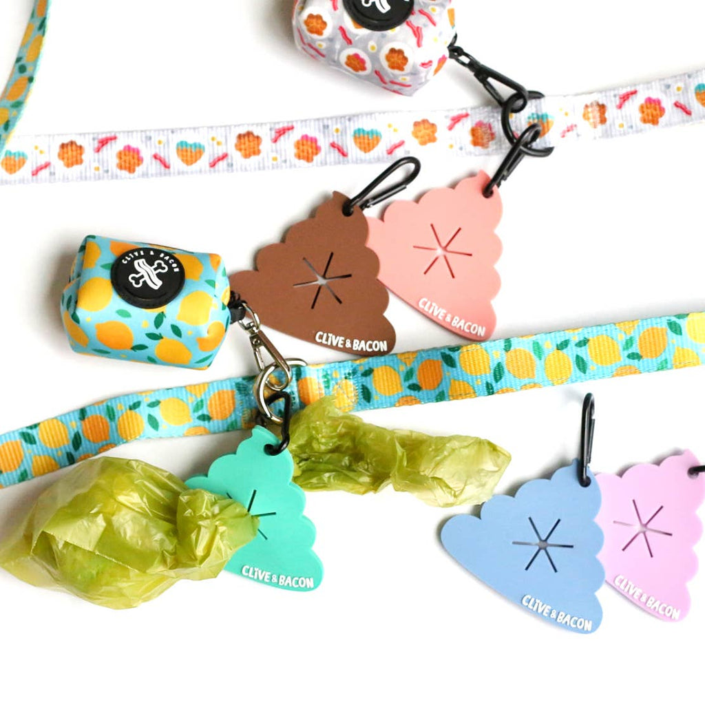 A set of cute dog poop-shaped bag dispenser keychains and matching patterned ribbon. The keychains come in brown, pink, green, and blue, with one keychain holding a lime green plastic bag.