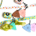 A set of cute dog poop-shaped bag dispenser keychains and matching patterned ribbon. The keychains come in brown, pink, green, and blue, with one keychain holding a lime green plastic bag.